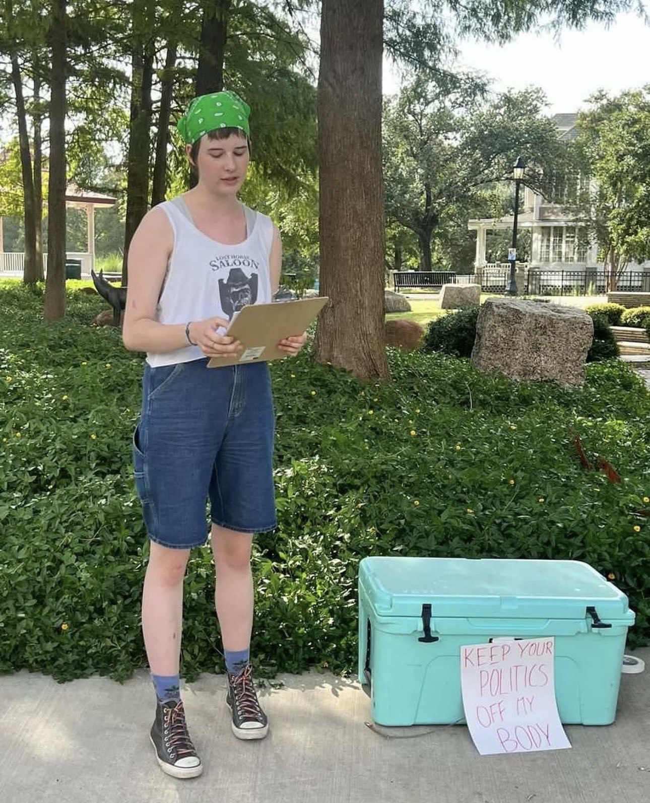 Louise Culbertson, a white teenage woman is holding a clipboard, standing next to a blue cooler with a sign on it reading "Keep your politics off my body."