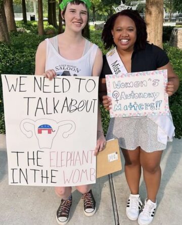 A close-up of Louise Culbertson, left, a white teenager posing with a young Black woman, each holding protest signs and smiling as they lean close together for the photo. Culbertson's sign reads "We need to talk about the elephant in the womb" with a drawing of a uterus with a GOP logo inside. The other sign rerads "Women's Autonomy Matters!"