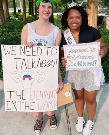 A close-up of Louise Culbertson, left, a white teenager posing with a young Black woman, each holding protest signs and smiling as they lean close together for the photo. Culbertson's sign reads "We need to talk about the elephant in the womb" with a drawing of a uterus with a GOP logo inside. The other sign rerads "Women's Autonomy Matters!"