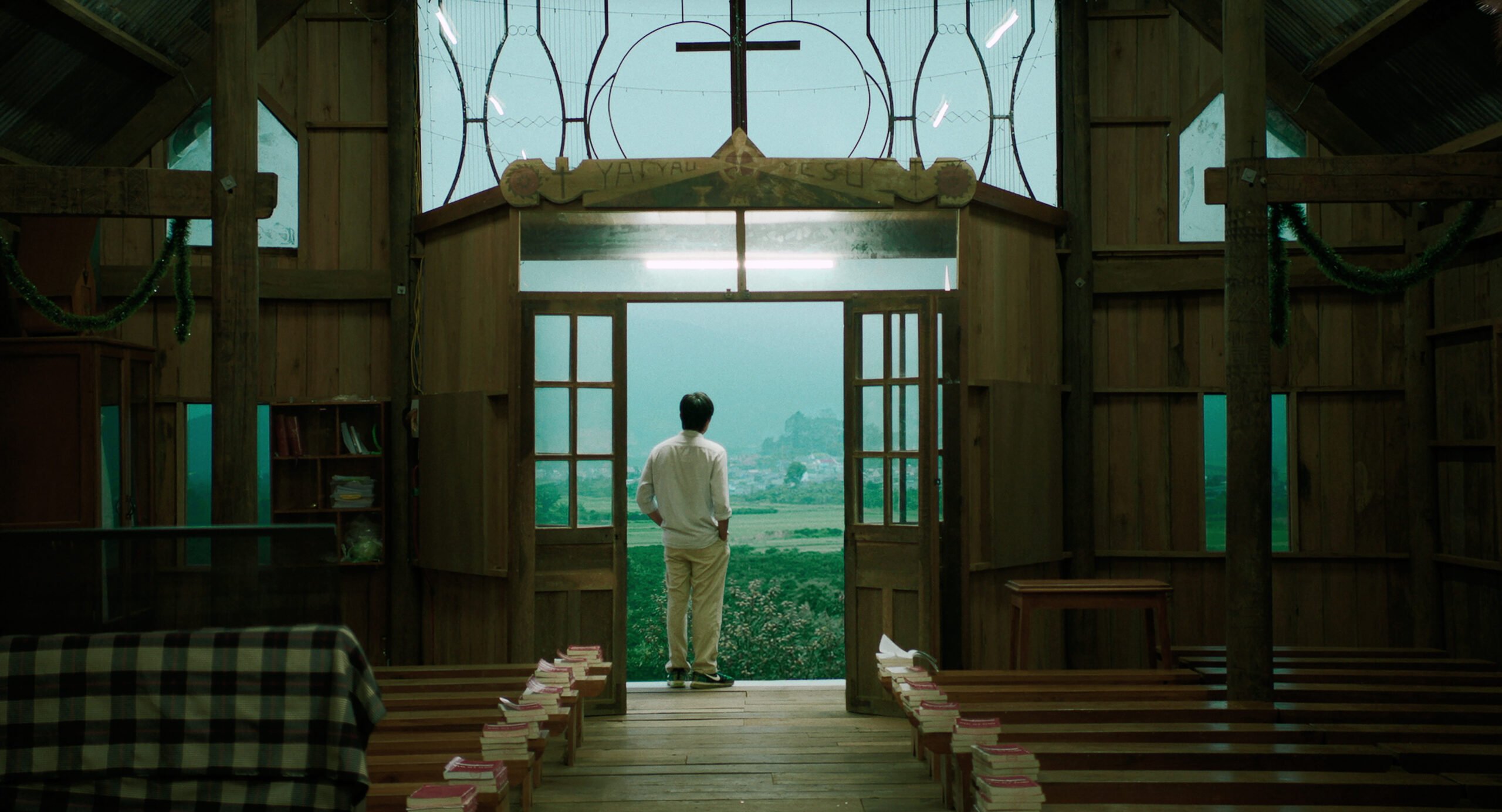 A lone figure in a white clothing, seen from behind, stands artfully framed in the glass doorway of an old-fashioned. but well cared for church in the rural countryside.