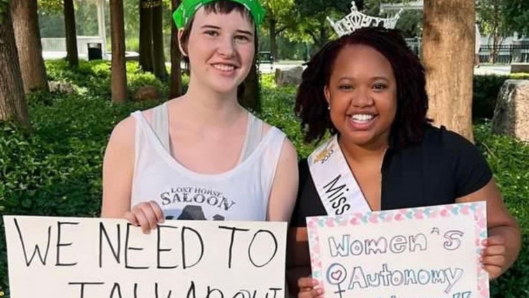 A close-up of Louise Culbertson, left, a white teenager posing with a young Black woman, each holding protest signs and smiling as they lean close together for the photo.