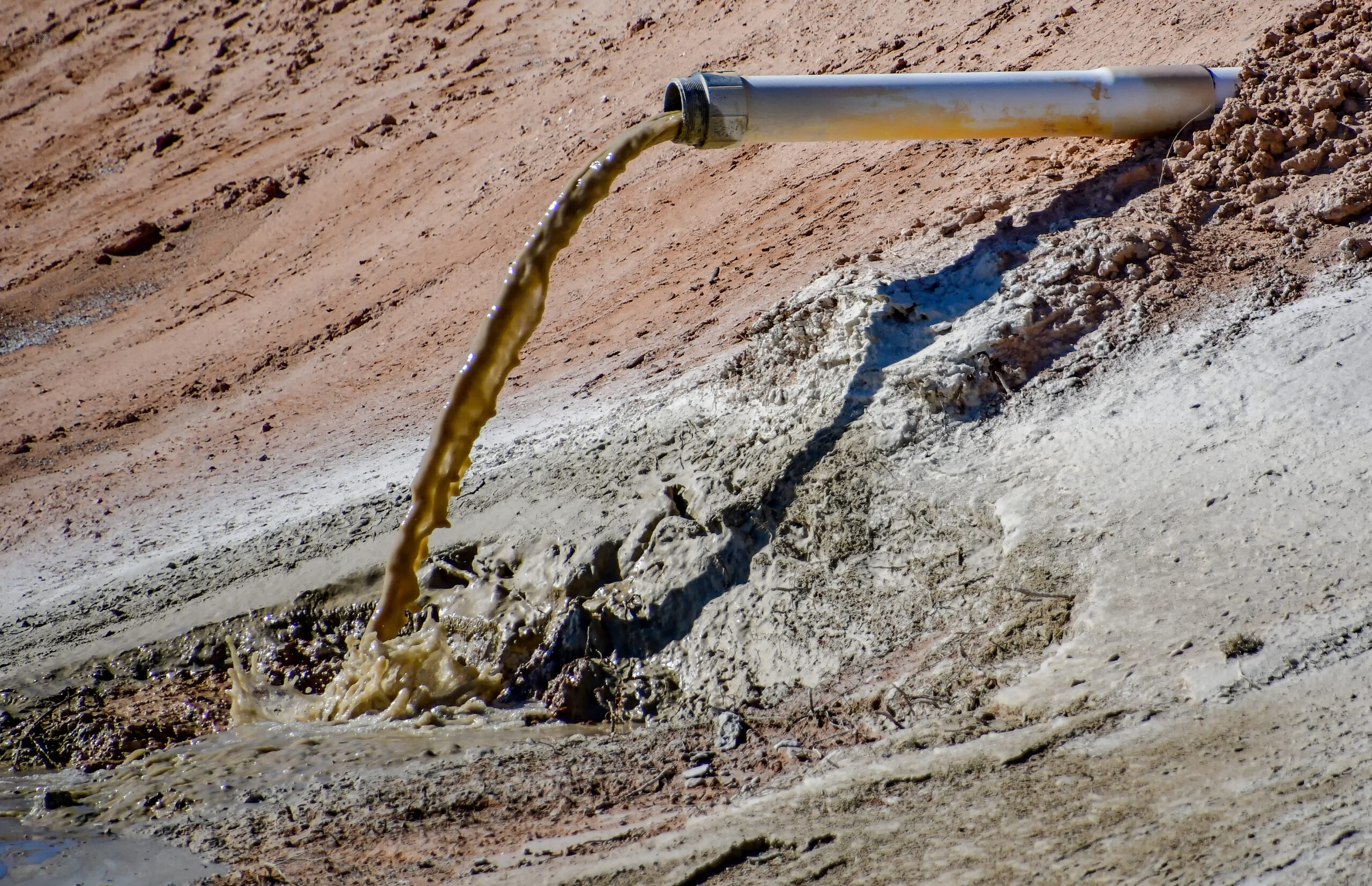 A stream of brown liquid spills from a long pipe onto a polluted looking, muddy and desolate field.