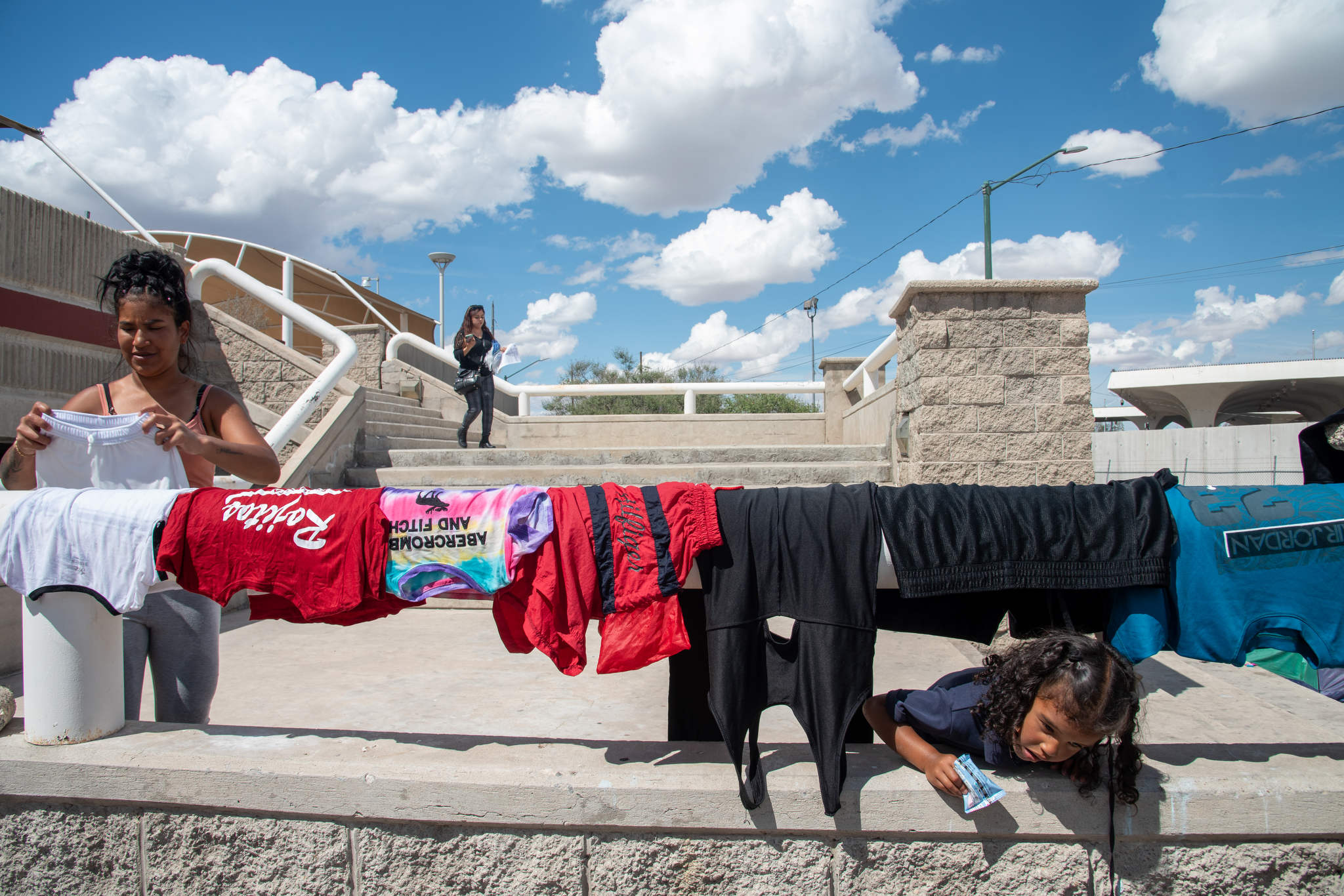T-shirts and other clothing hang on a metal railing of a municipal building, under a partly cloudy sky. The daughter is playfully looking under the railing while holding a snack.