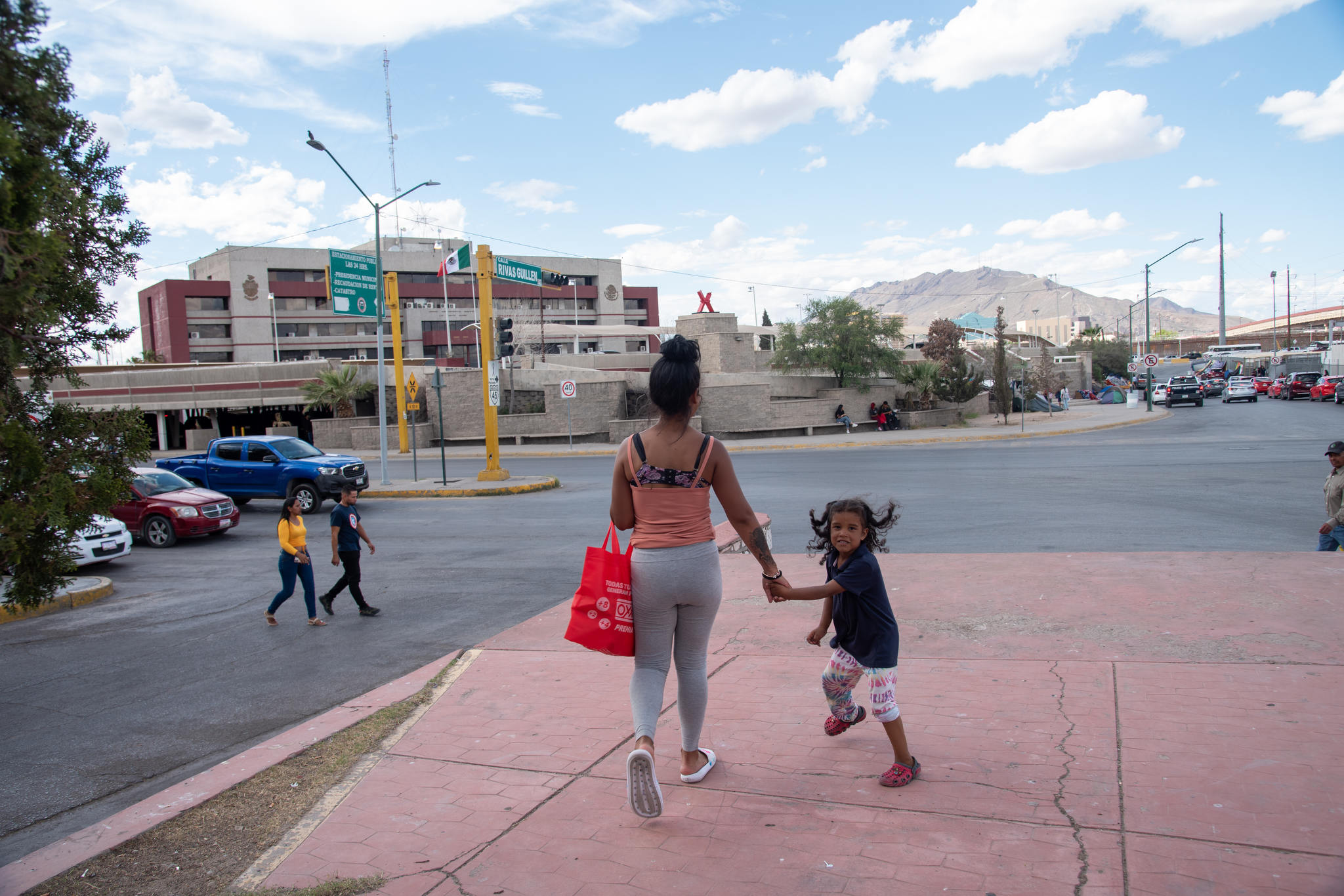 A Latinx woman walks with her back to the camera, holding her young daughter's hand, cwho is looking back at us as she energetically walks with mom down an open urban street.