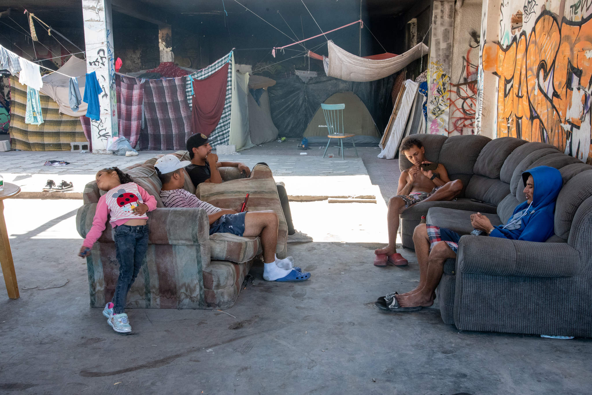 A group of people sit, some shirtless, using their phones on dirty abandoned couches in the graffiti-covered ruins they call their temporary home. Rooms are made with hanging fabric in the background.