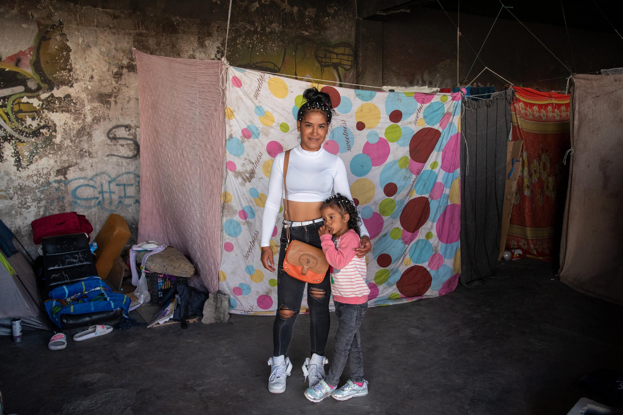 A Latinx mother and her waist-high daughter stand embracing by their home, a cement, graffiti covered ruin separated into makeshift shelters using hanging fabric.