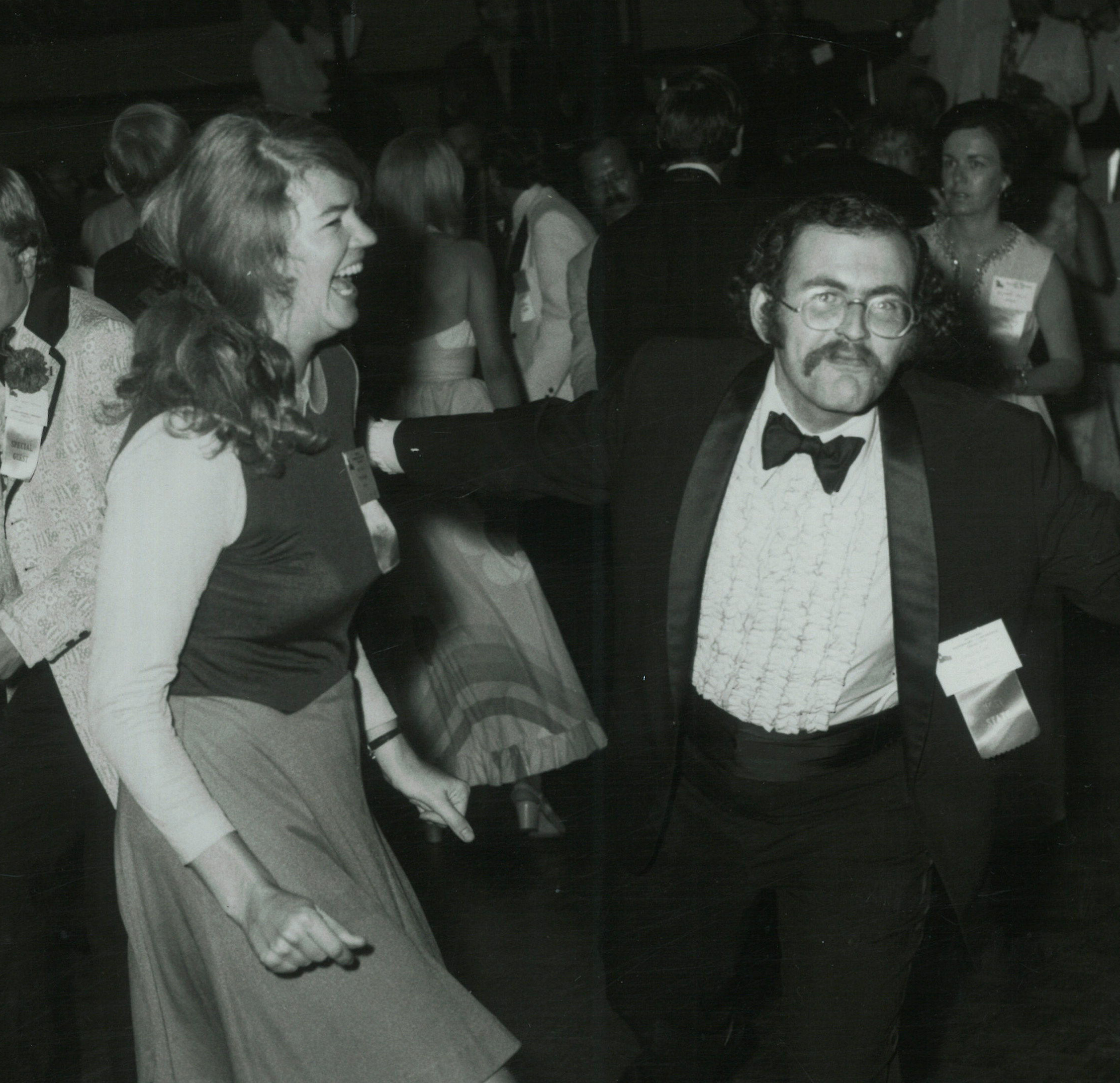 In a black and white photo, Molly Ivins, wearing a skirt and sleeveless shirt over a white blouse, laughs as Carlton Carl, in a tuxedo and bow tie, gestures dramatically next to her.