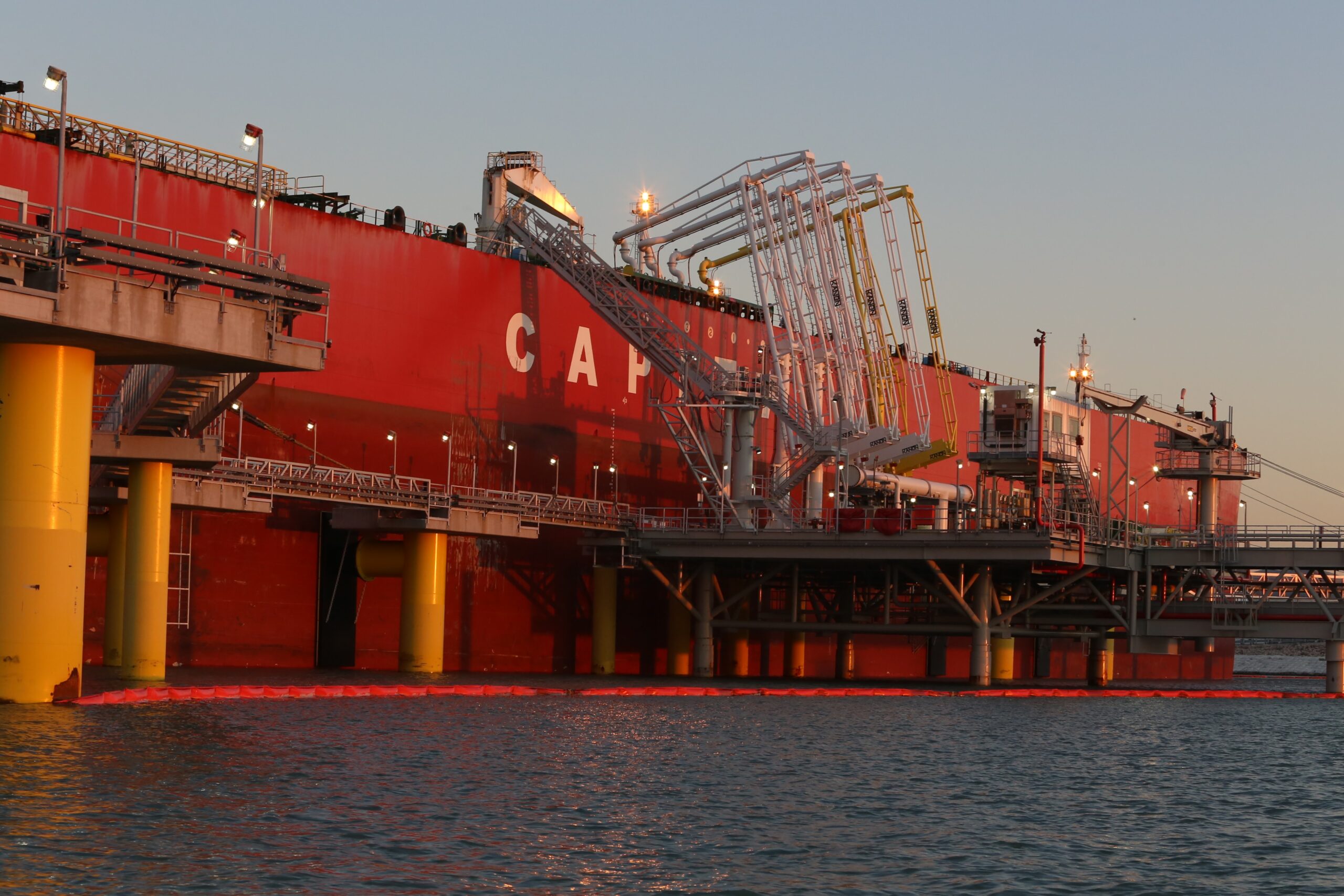 Tall piping and a gangway attach a massive red tanker to a dock.