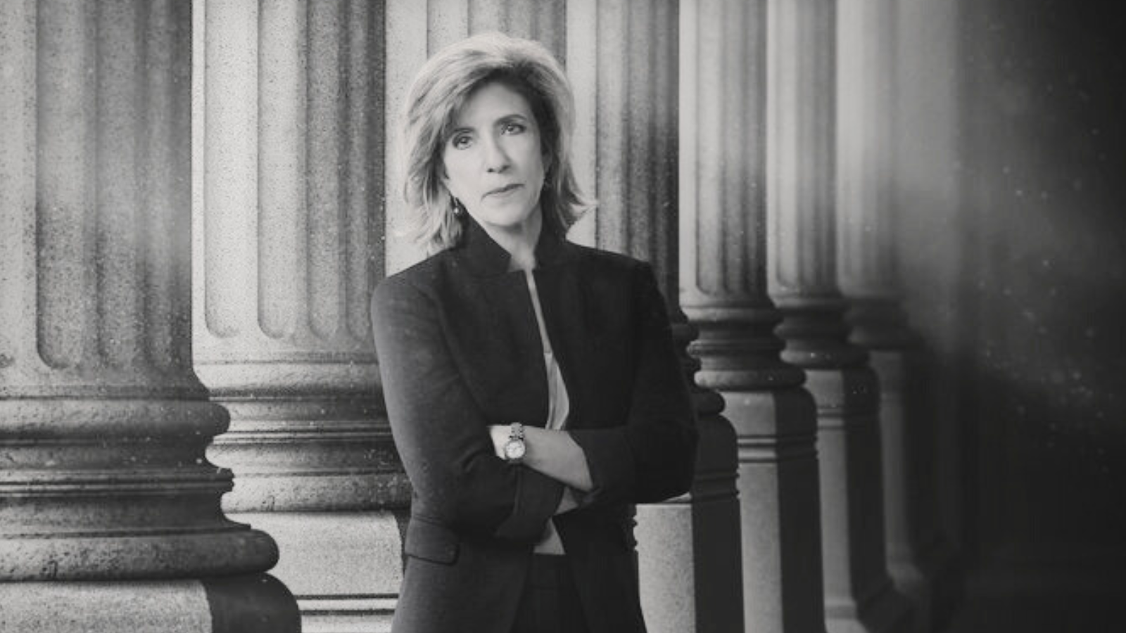 Kelly Siegler, in a suit jacket and pants, stands with her arms crossed in front of columns similar to the kind found outside courthouses. A dramatic black and white film filter was applied to the photo.