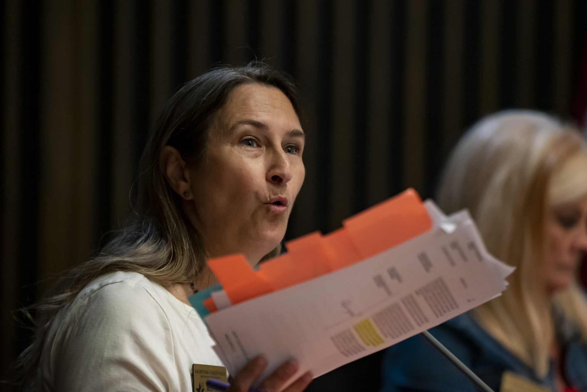 A dark-haired woman gestures with a handful of paperwork as she speaks.