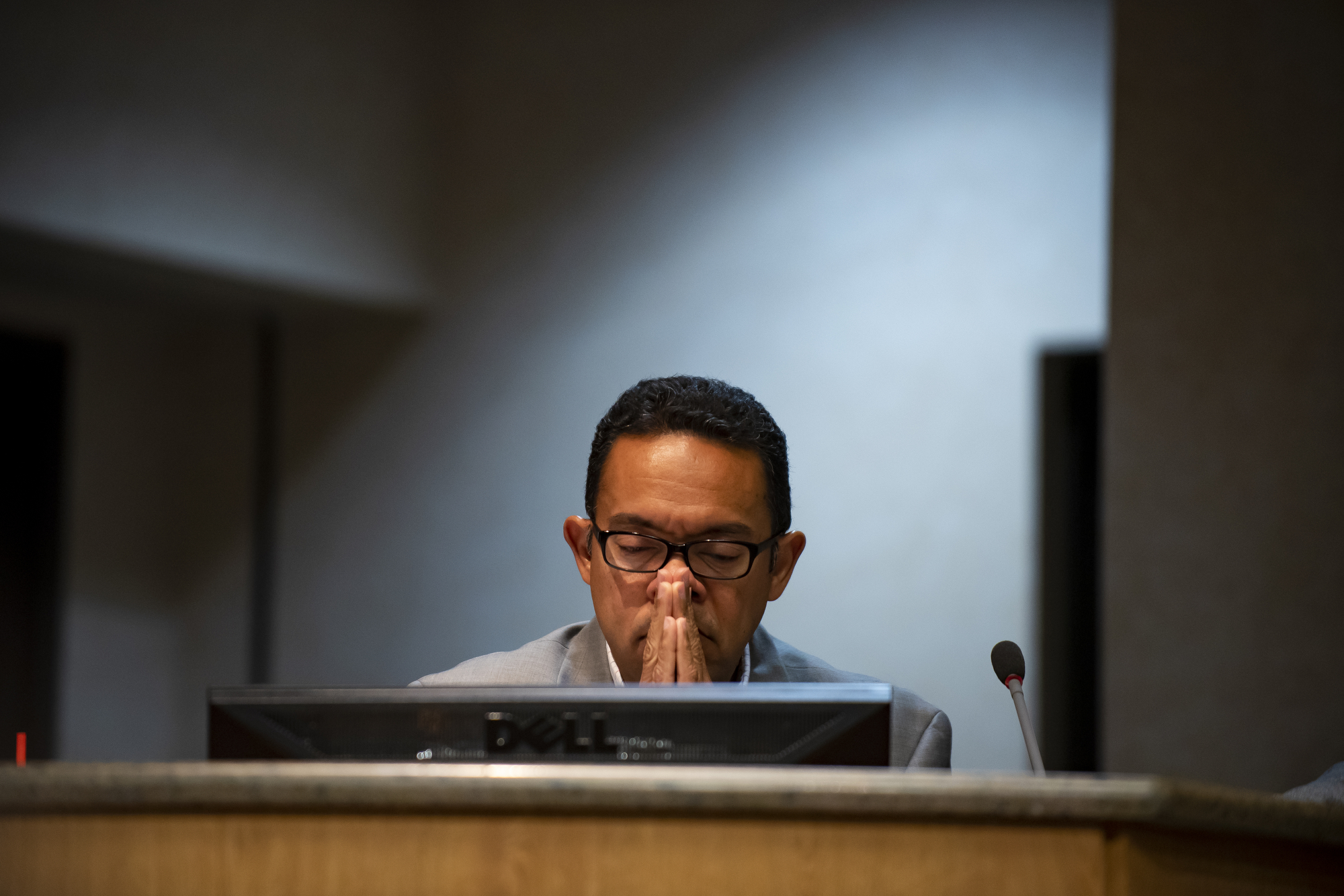 A short-haired Latinx man folds his hands and closes his eyes as if frustrated.