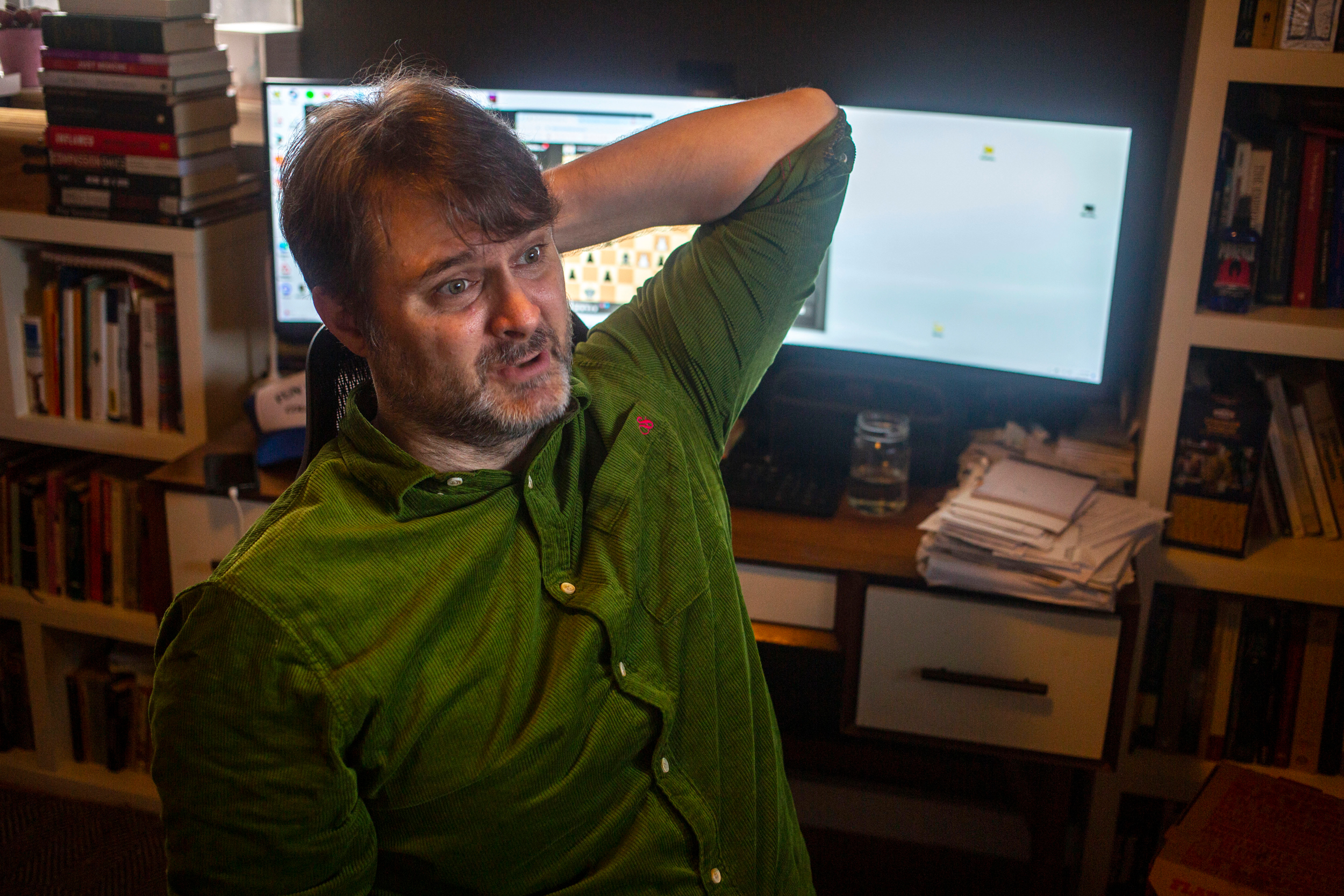 Gabriel Clark-Leach, wearing a green button down, sits in a relaxed pose in front of his computer in his office, one hand behind his head.