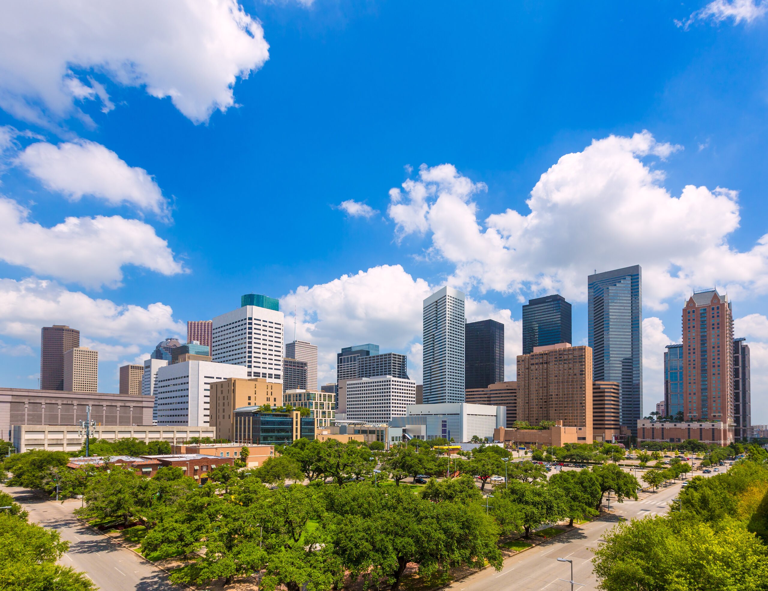 The Houston skyline, with its numerous recognizable skyscrapers, seen from a greenbelt to the south, under a parly cloudy blue sky.