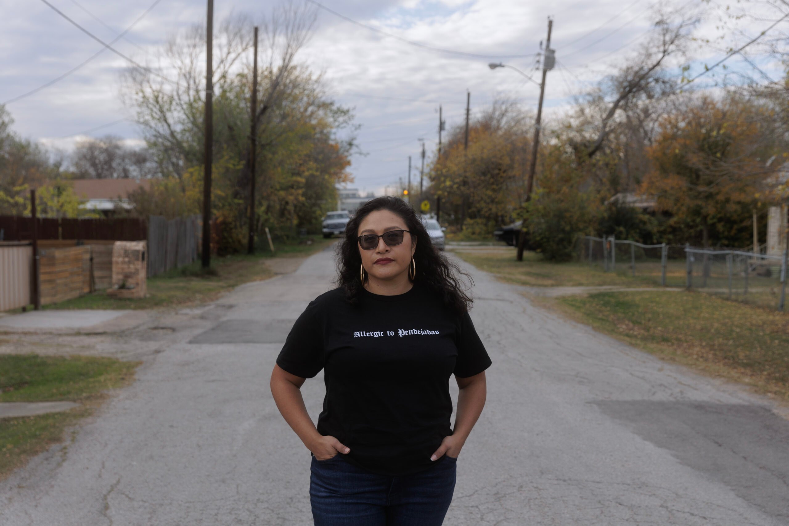 A latinx woman in a black t-shirt reading "Allergic to Pendebrajas" stands in her suburban neighborhjood in West Dallas.