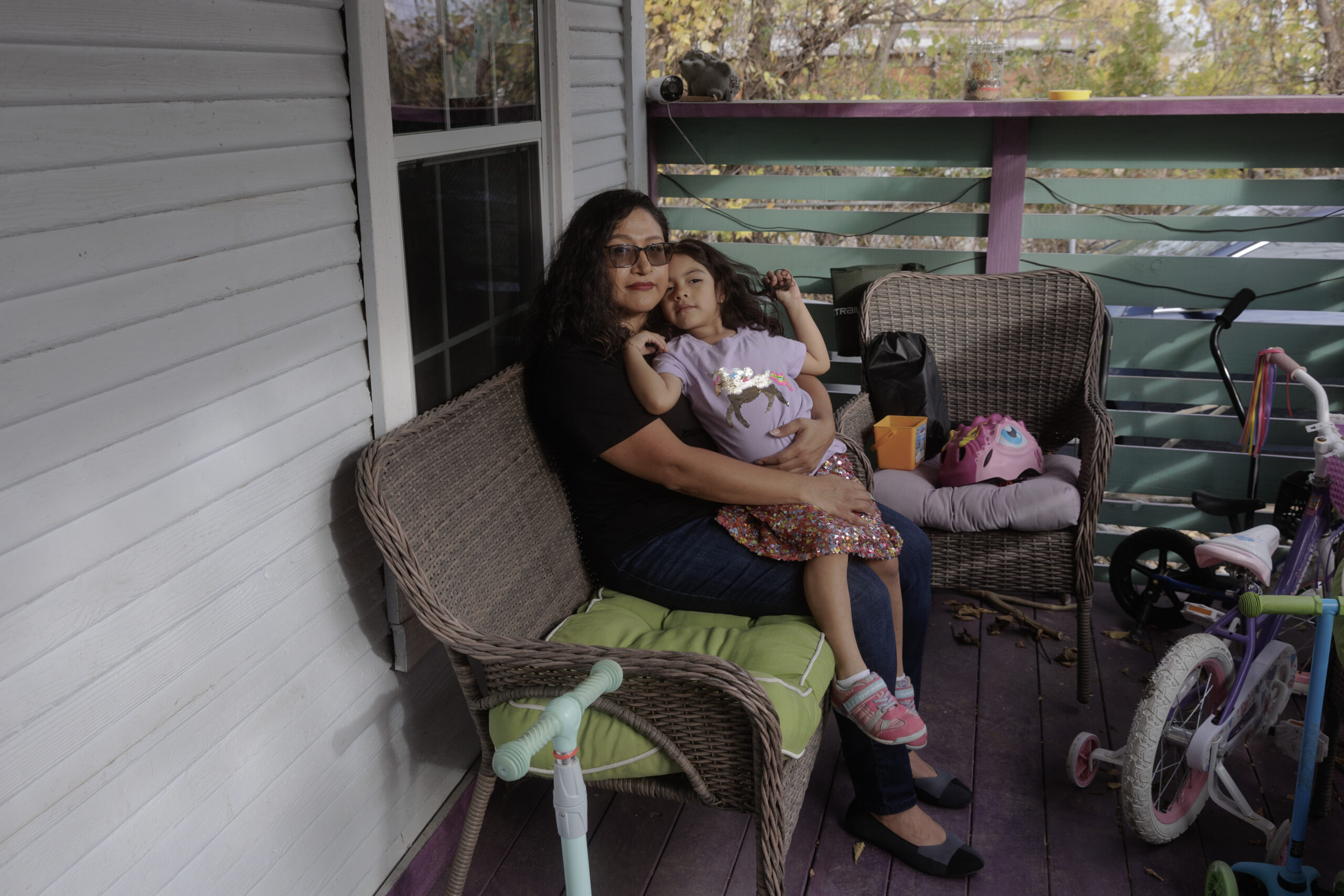 Sitting on a comfortably cluttered porch, a Latinx woman holds her daughter in her lap.