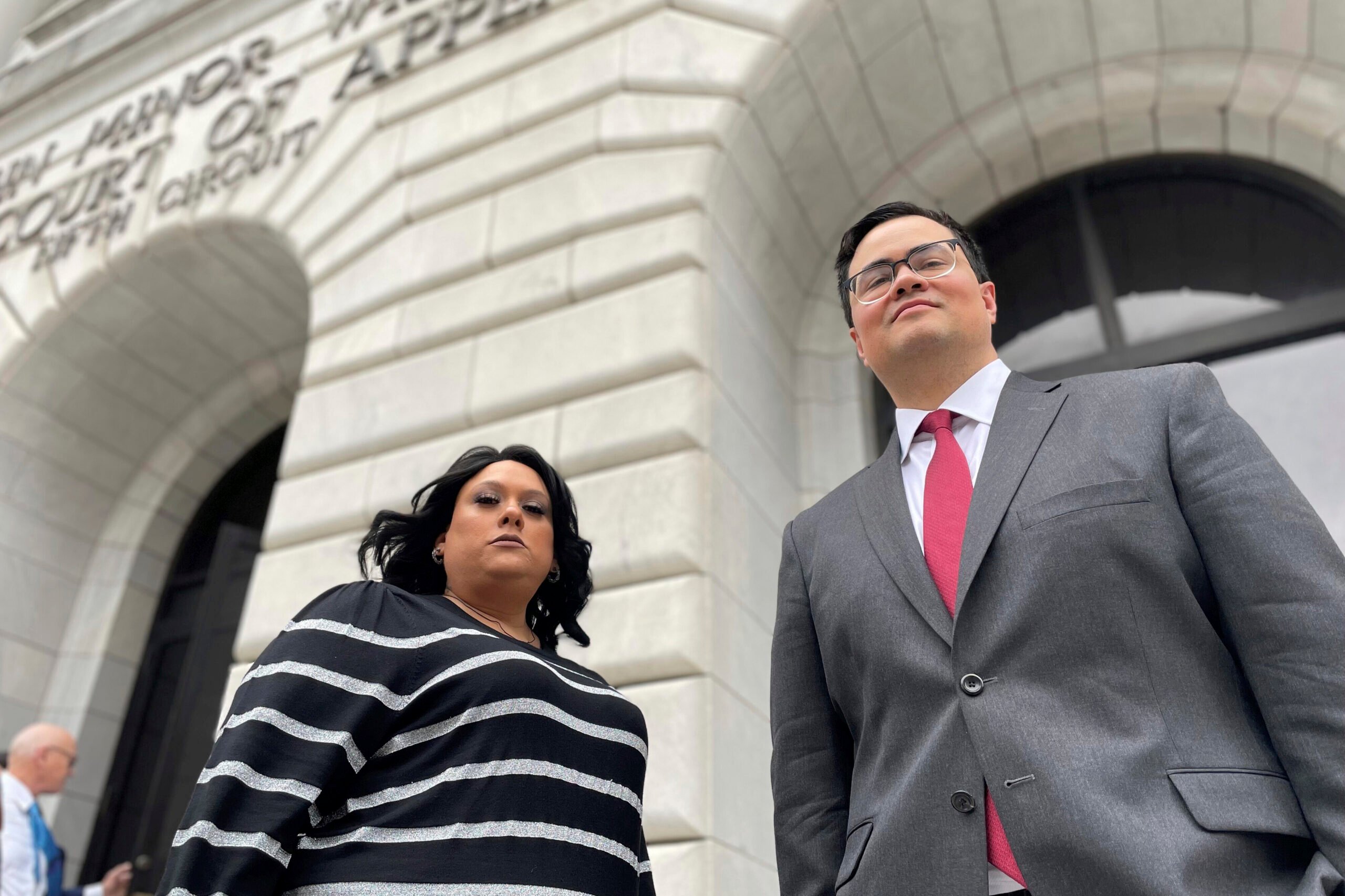 A Latino woman and man stand with serious, proud expressions outside of a courthouse. Priscilla Villarreal is in a black and white striped sweater while her lawyer, J.T. Morris, wears a suit and tie.