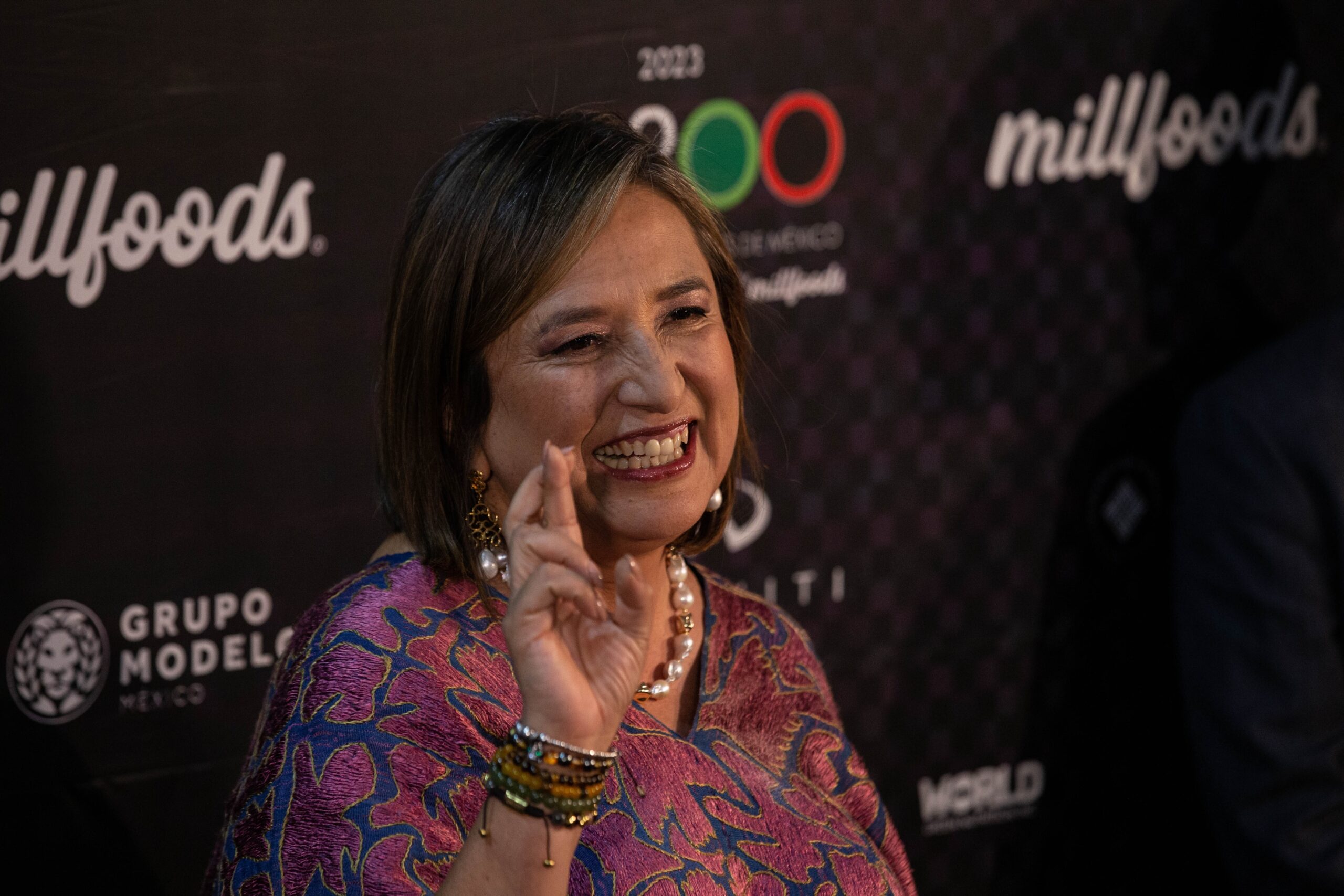 Bertha Xóchitl Gálvez Ruiz, a candidate in the upcoming election in Mexico, is standing in front of a step-and-repeat, holding up her hand and smiling.