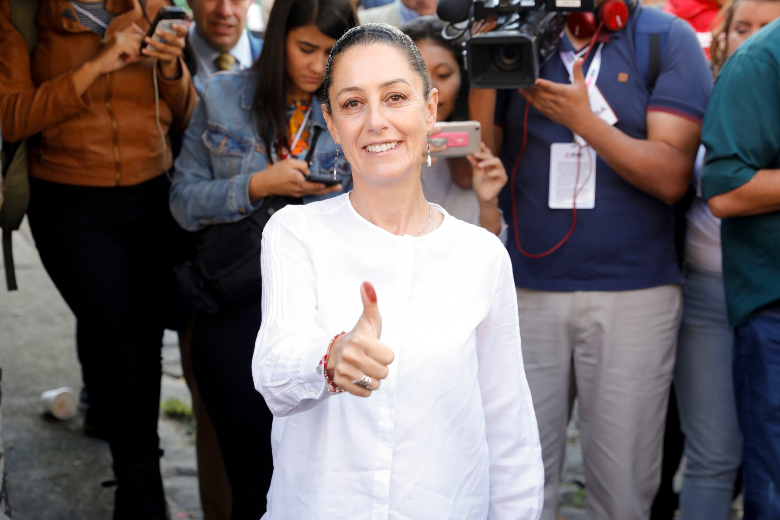 Claudia Sheinbaum Pardo, a Latinx woman in a white dress, gives a thumbs up to the camera, showing the ink on her thumb. Photojournalists crowd behind her with their cameras.
