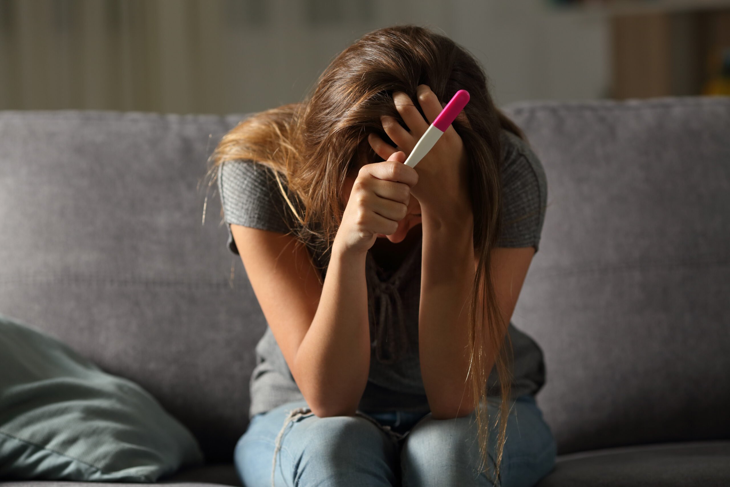 A young woman or teen sitting on a couch, head in hands with sadness or despair, as she holds a pregnancy test in one hand.