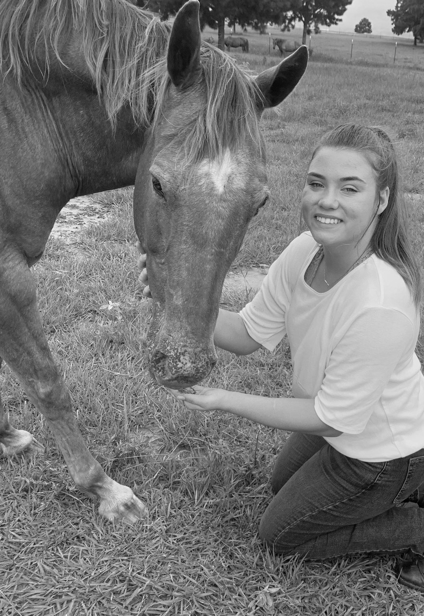 A young woman with brown hair kneels next to a horse. A black and white filter is applied to the photo.