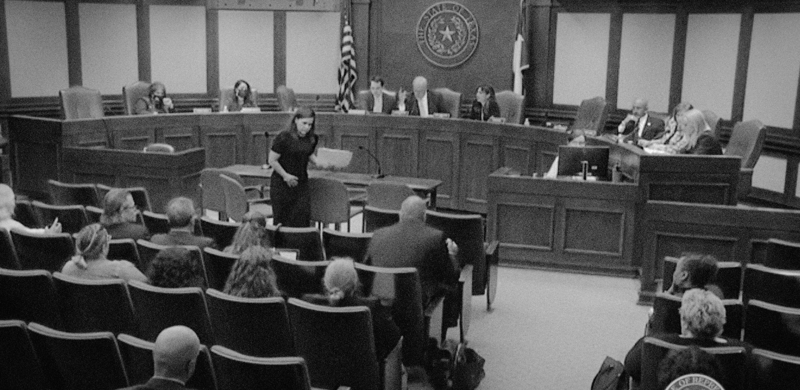 In a black and white screenshot from a live feed, a white woman holding a manila folder testifies in front of a committee in the Texas House of Representatives.