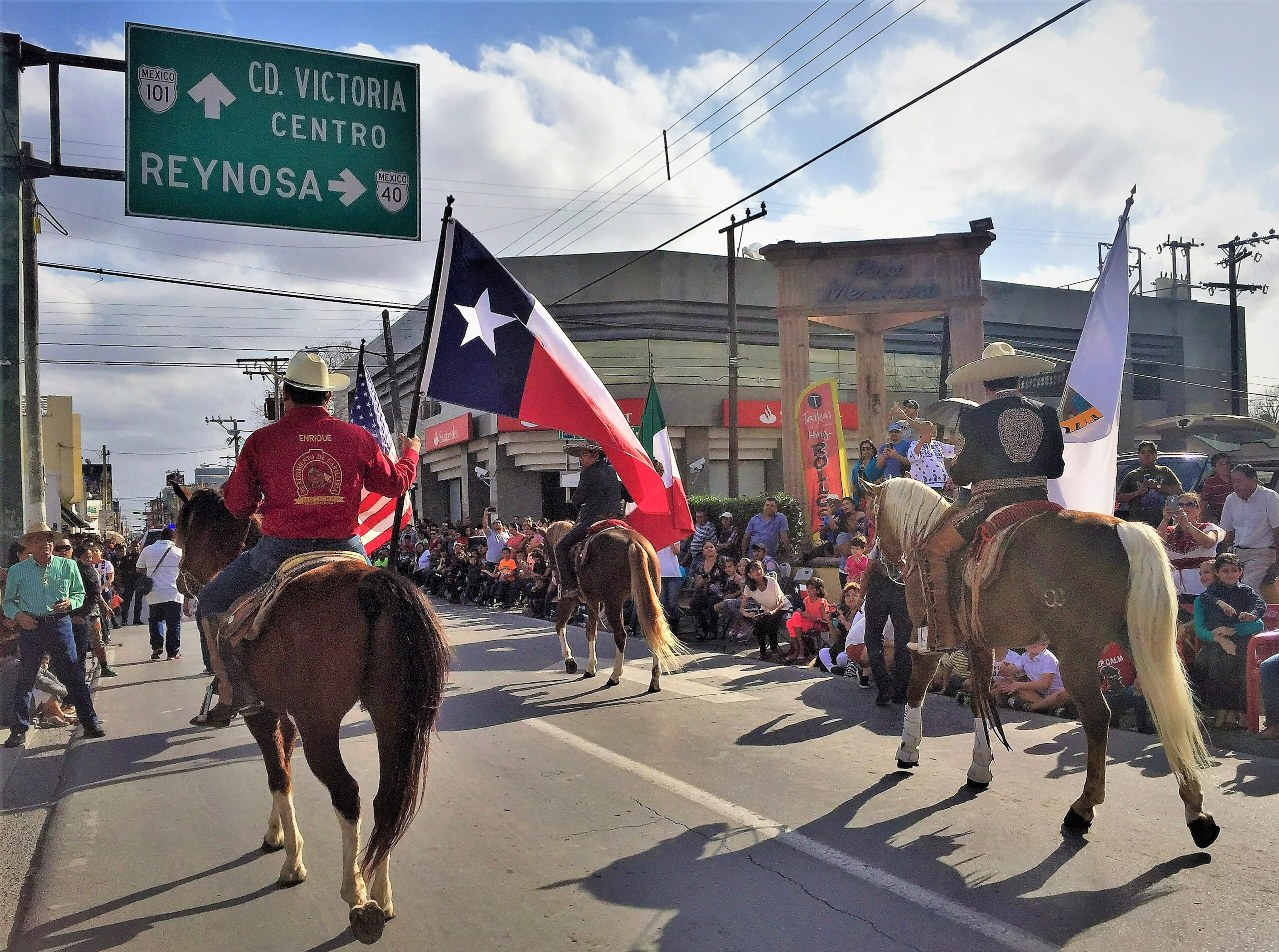 A parade along the border (frontera) with participants in cowbow hats on horseback. The main rider in focus is holding a Texas flag. Viewers cheer from along the edge of the street in a dense crowd.