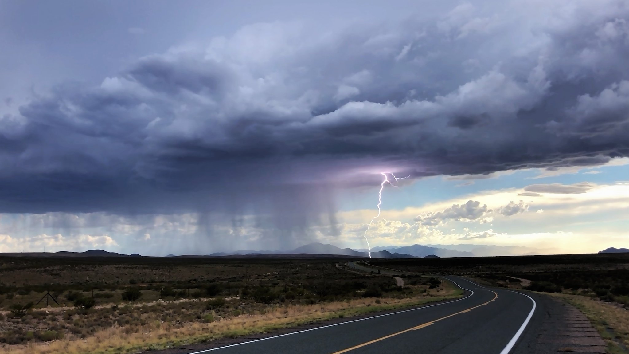 A wide-angled photo of a dramatic stormcloud over a small rural highway. A bolt of lightning shoots from the cloud in the distance, where intense rain can also be seen.