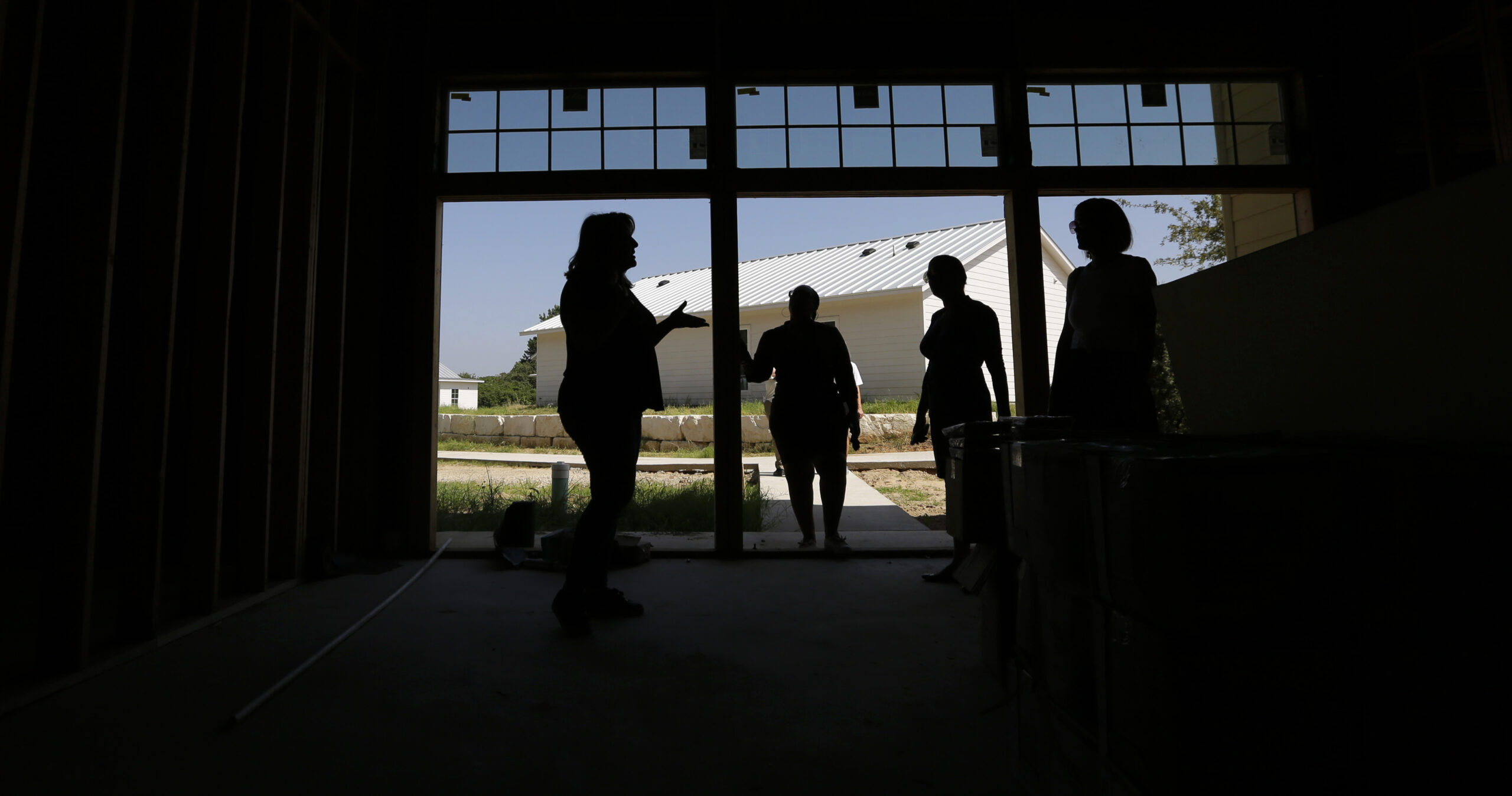 Four silhouetted figures stand under a wood and glass awning on a ranch, with a white barn visible in the distance.