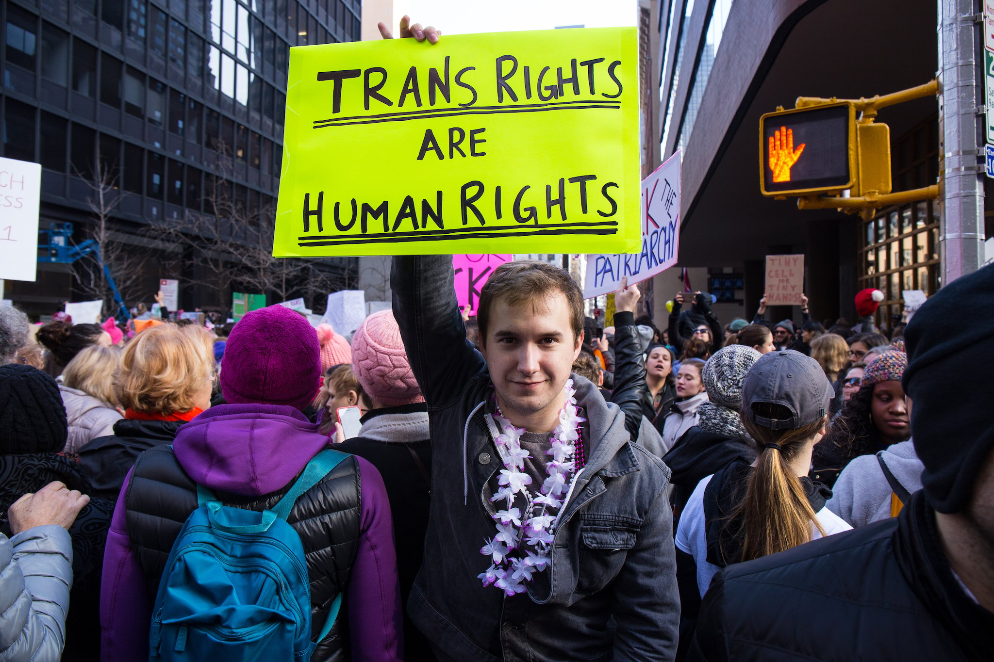 A man in a crowded, urban protest march turns away from the marchers to face the camera, smiling as he holds up a handwritten sign reading "Trans Rights are Human Rights."