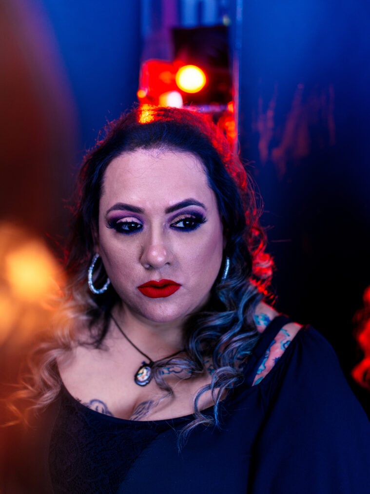 A dramatic close up of Priscilla Villarreal, wearing a scoop neck black tee, a cameo necklace, large silver hoop earrings and bold makeup.