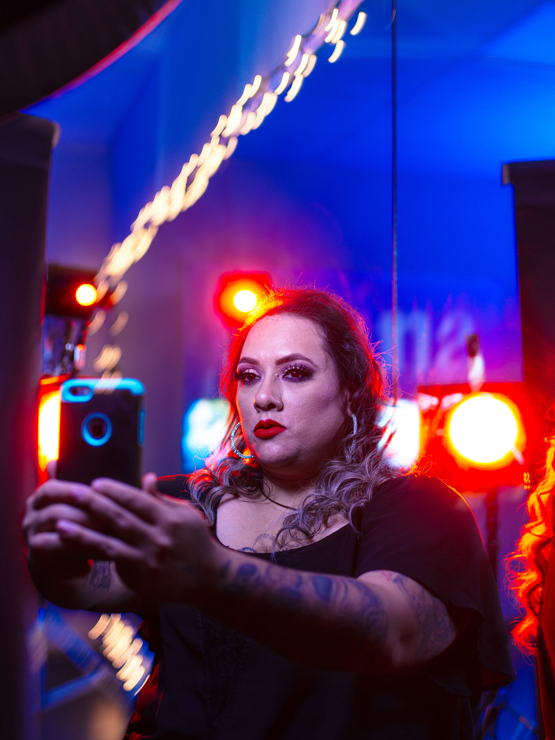 In a moodily lit, starkly colored night time photograph, Priscilla Villarreal takes a selfie on her iPhone. She's a Latino woman in a black scoop neck shirt, with wave hair with blond highlights, wearing bold red lipstick and eye liner.