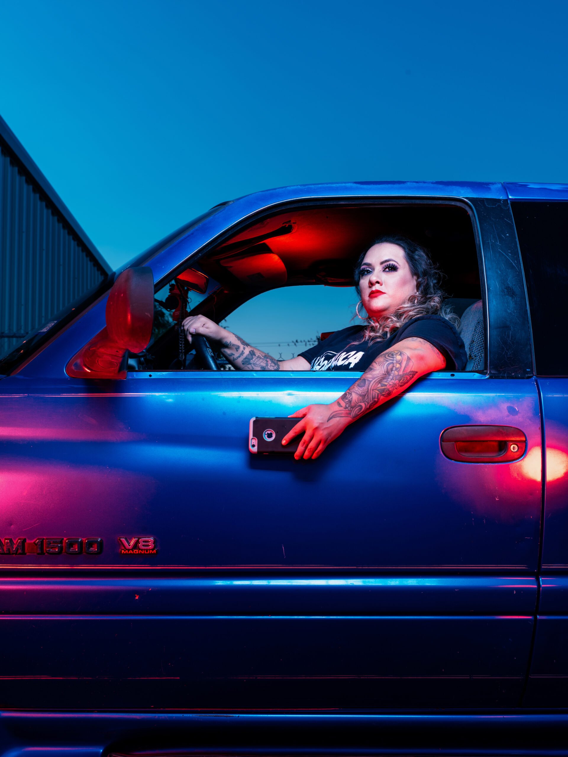 Priscilla Villarreal, or La Gordiloca, a Latino woman, holds a smartphone in her hand out the window of her car. The photo is filtered to create sharp edges and starkly contrasting, deep colors. Villarreal is visible from the waist up, in a black graphic tee, wearing red lipstick and bold makeup, with extensive tattoos on her arms.