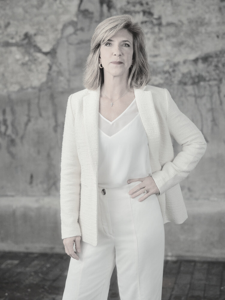 Kelly Siegler in a white suit with a v-neck shirt undenreath. She is standing with a hand on her hip. A black and white filter was applied to the photo.