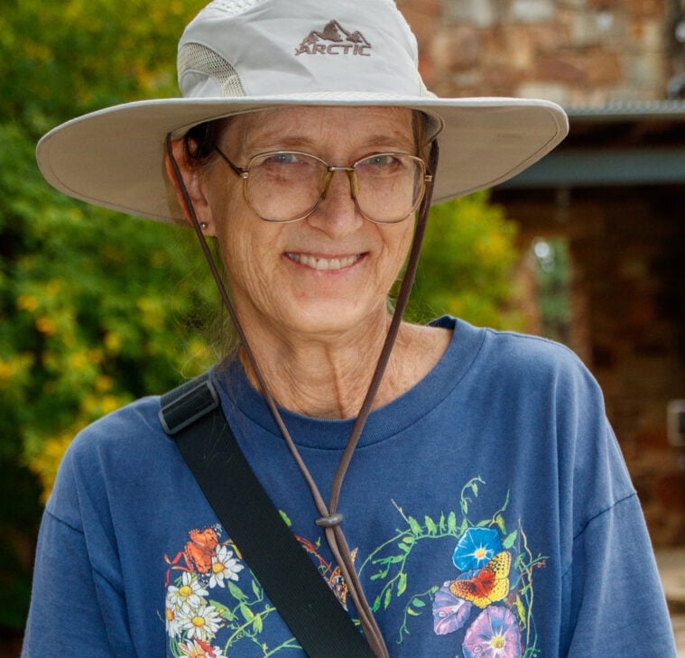 Valerie Bugh, an older white woman in a outdoor hat, wearing a floral blue t-shirt with a shoulder bag, smiles for the camera.