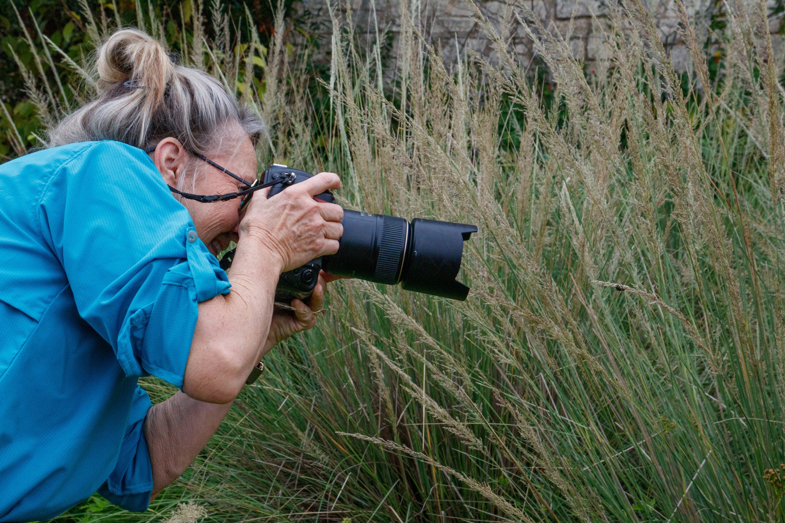 A white woman with graying hair, in a blue shirt, takes a photo with a long macro lens, focusing on an insect in the overgrown grass.