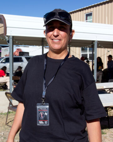 A woman in a black ball cap and black t-shirt with a homemade press pass in a lanyard holder. She's standing outside a ranch with people gathered behind her in a shade structure.