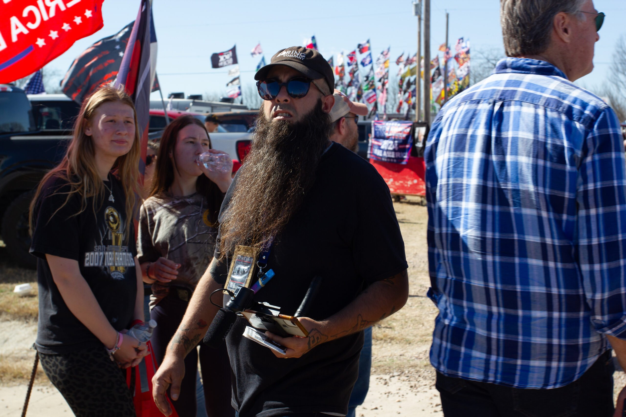 Jerry J. Pena-Ahuyon, a man with a very long dark beard, wearing sugnglasses and dark clothing, along with a digital SLR and homemade press badge around his neck.