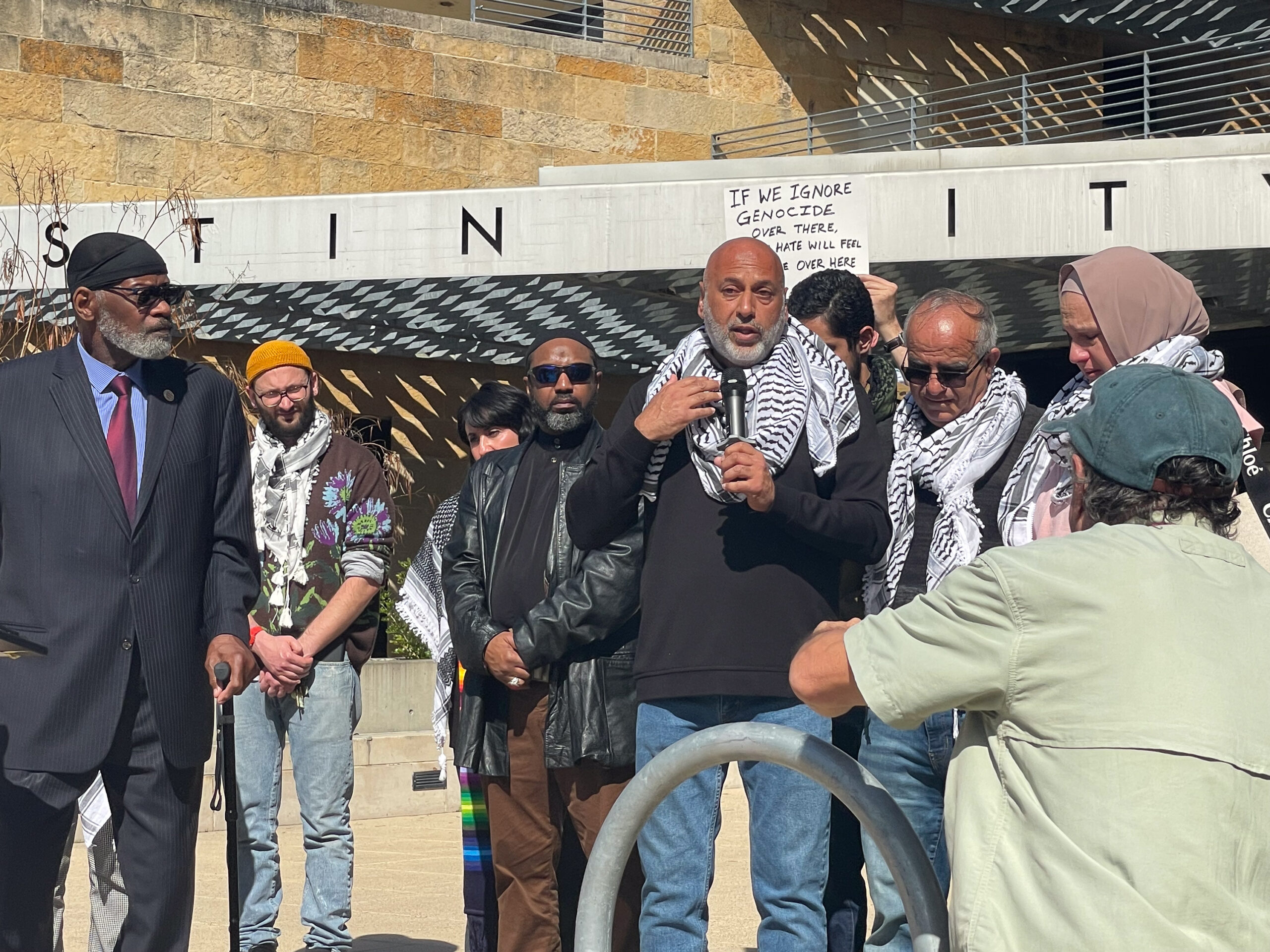 A man in a keffiyeh speaks emotionally into a microphone, surrounded by a diverse group of supporters, many of whom also wear Keffiyeh. They are standing directly outside the entrance to Austin City Hall.