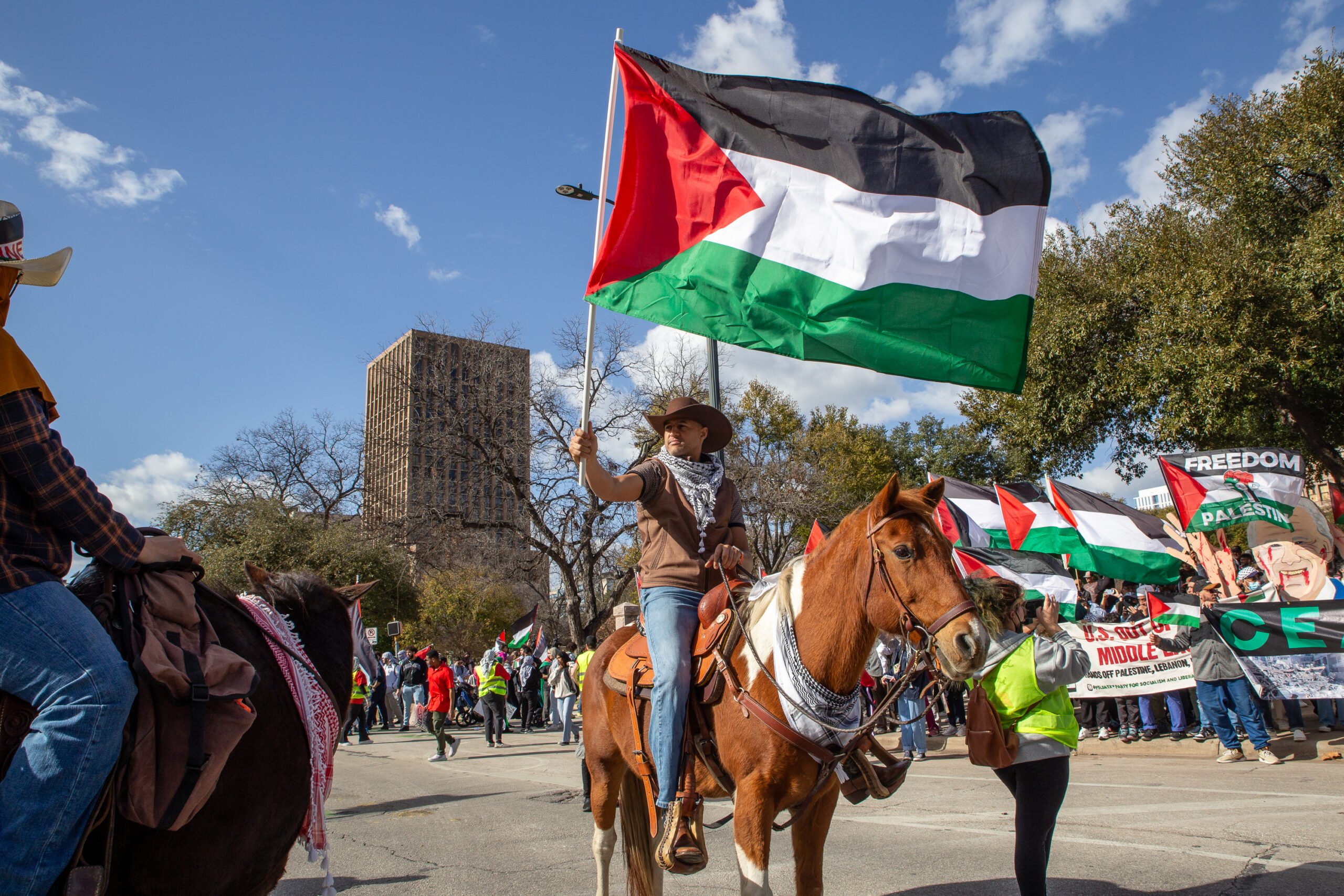 A man on horseback holds the Palestinian flag above his head, with a pro-Palestine demonstration in favor of a ceasefire resolution behind him. He's wearing Western clothing, with both the horse and ride wearing keffiyeh.