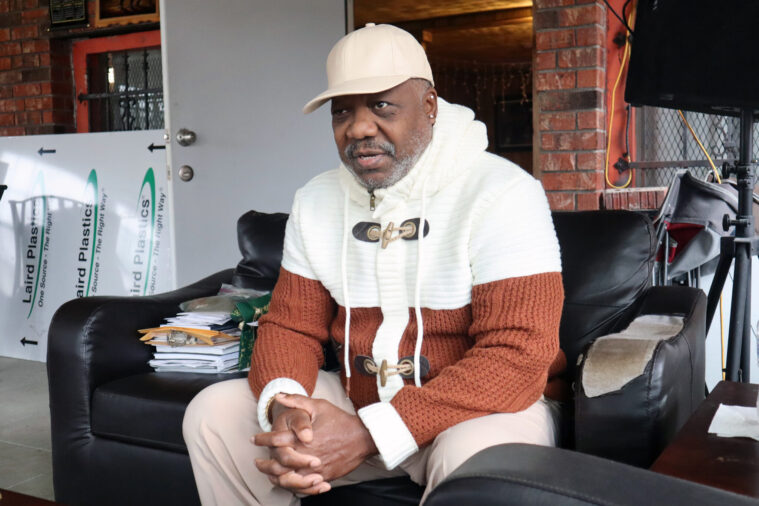 Hilton Kelley is a Black man with a light, graying beard and mustache, seated on a leather couch in a living roomring a type space. He's wearing an off-white ball cap with a white and brown colorblock hoodie. 