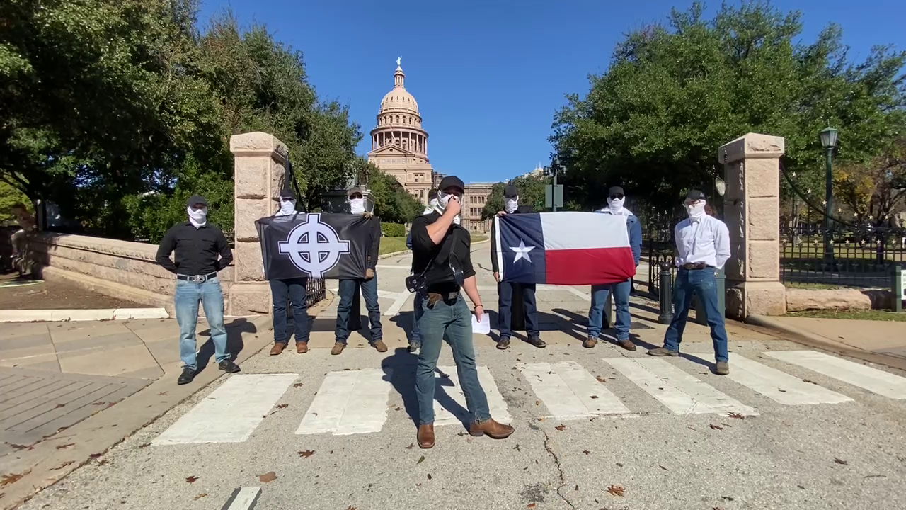 A group of masked protesters gather at the gate to the Texas Capitol with a Texas flag and a Celtic Cross banner.
