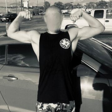 A white man in a black tank top with a Texas-based logo on the chest flexes his muscles. He is standing outside in a parking lot.