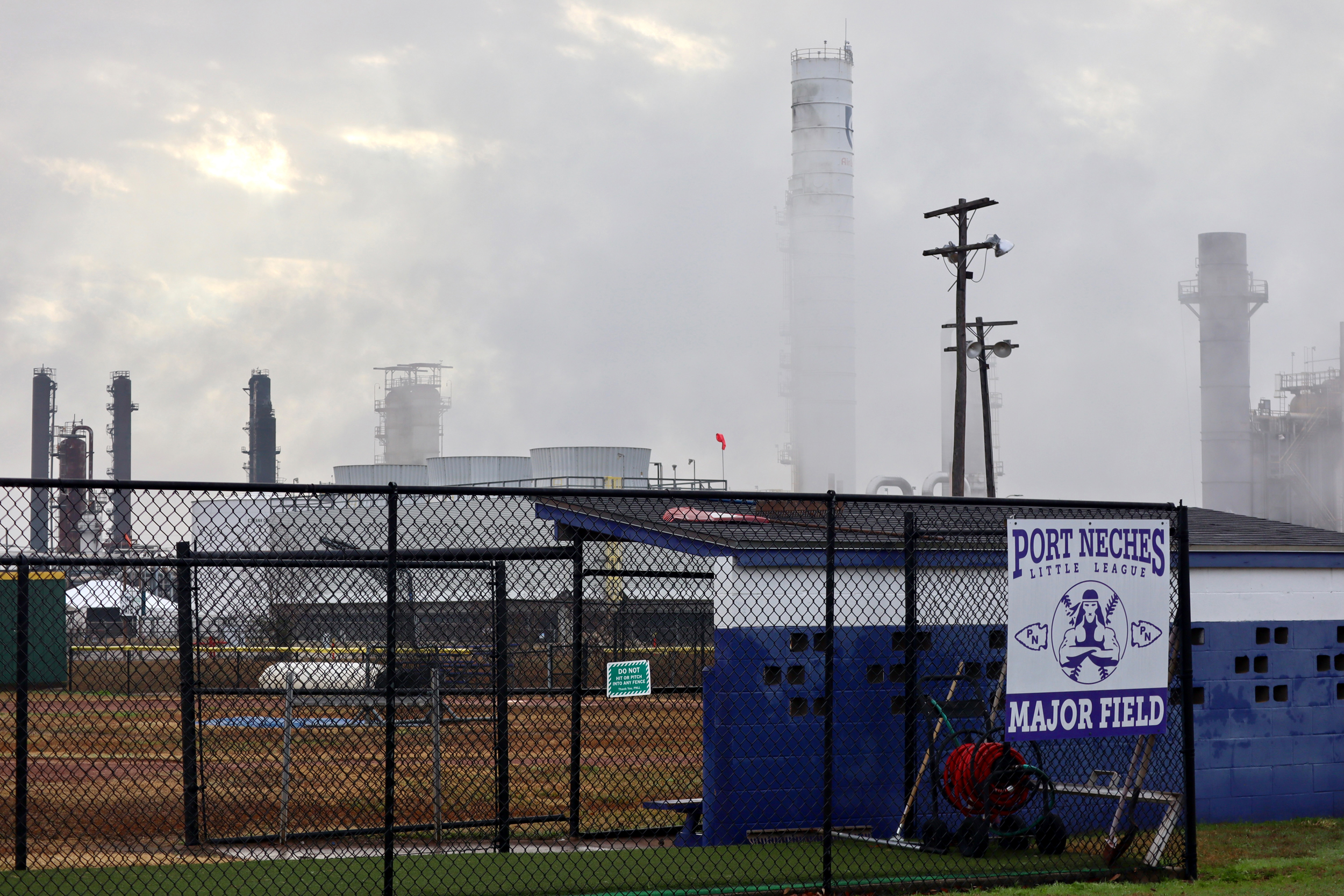 The steaming smokestacks of a petrochemical plant stand directly a Little League ball field.