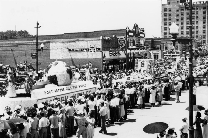 A black and white photo of a parade through downtown Port Arthur, featuring a crowd gathered around a float which reads "Port Arthur oils the world."